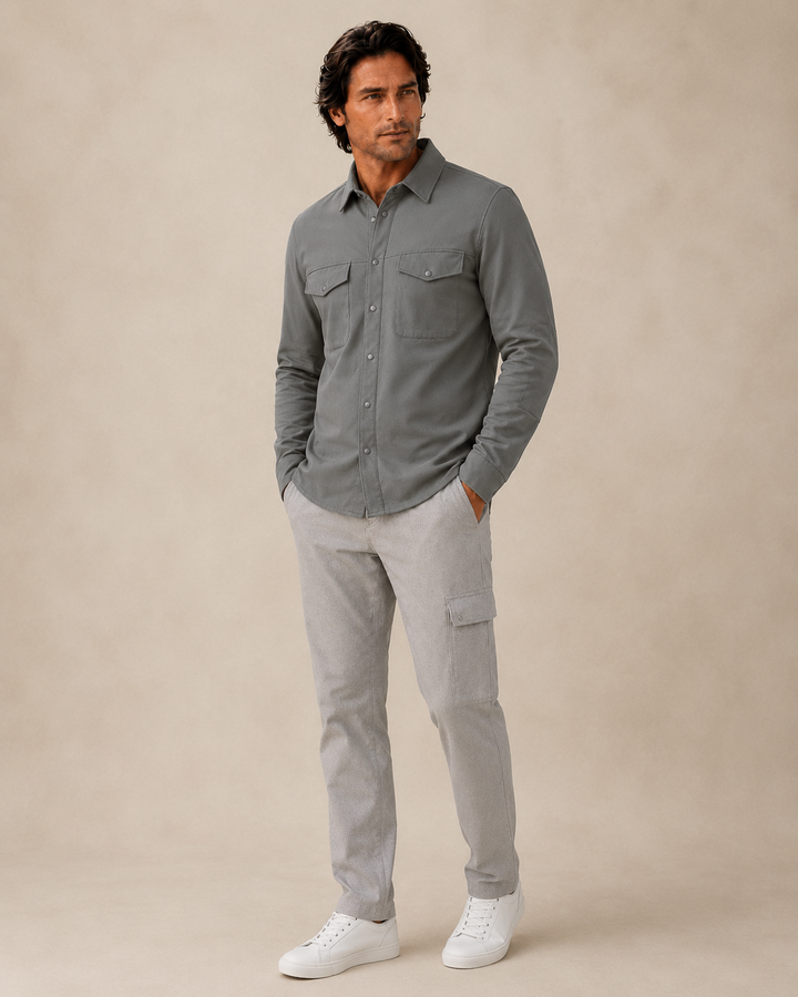 A man modeling a Buki Atlas Over Shirt button-up shirt with two chest pockets, paired with light gray Venture cargo pants and white sneakers, standing against a neutral beige studio backdrop.