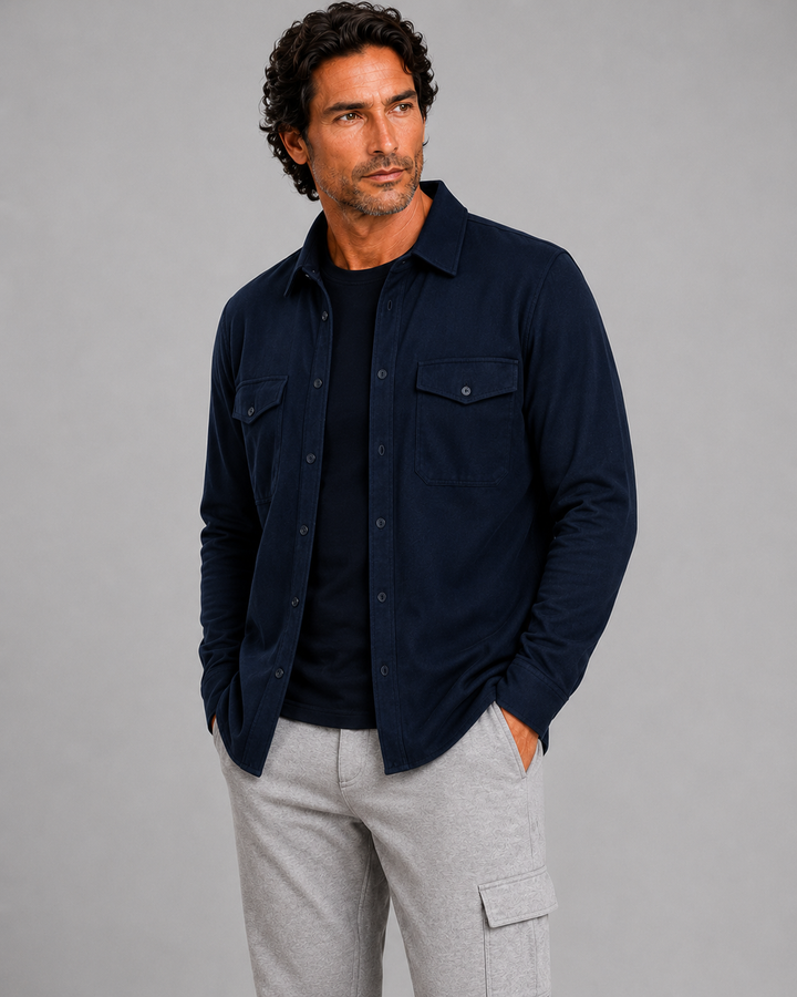 A man modeling a dark navy blue Atlas Overshirt over a black t-shirt, paired with light gray cargo pants and white sneakers, standing against a textured beige studio backdrop.