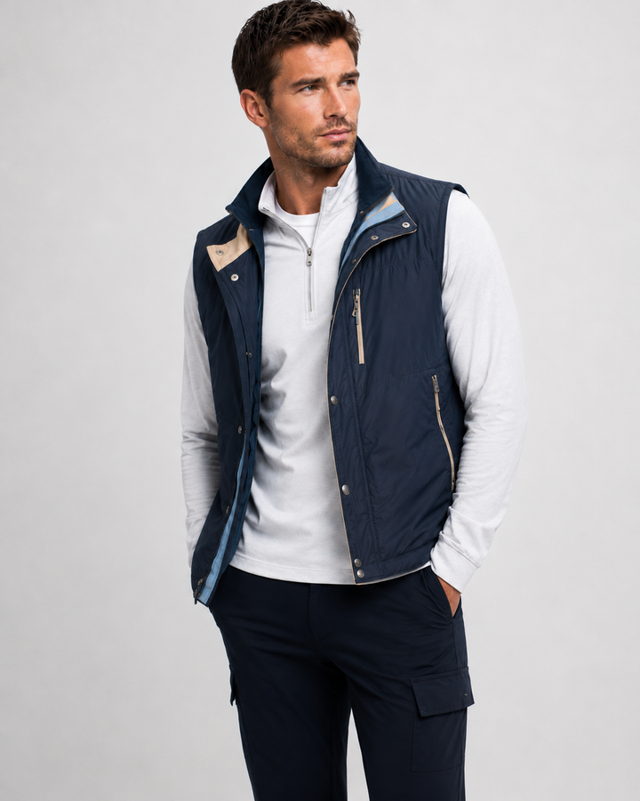 A man wearing a navy blue sleeveless quilted BUKI TECH vest over a white long-sleeve 1/2 zip  and navy pants, photographed against a light gray studio background.