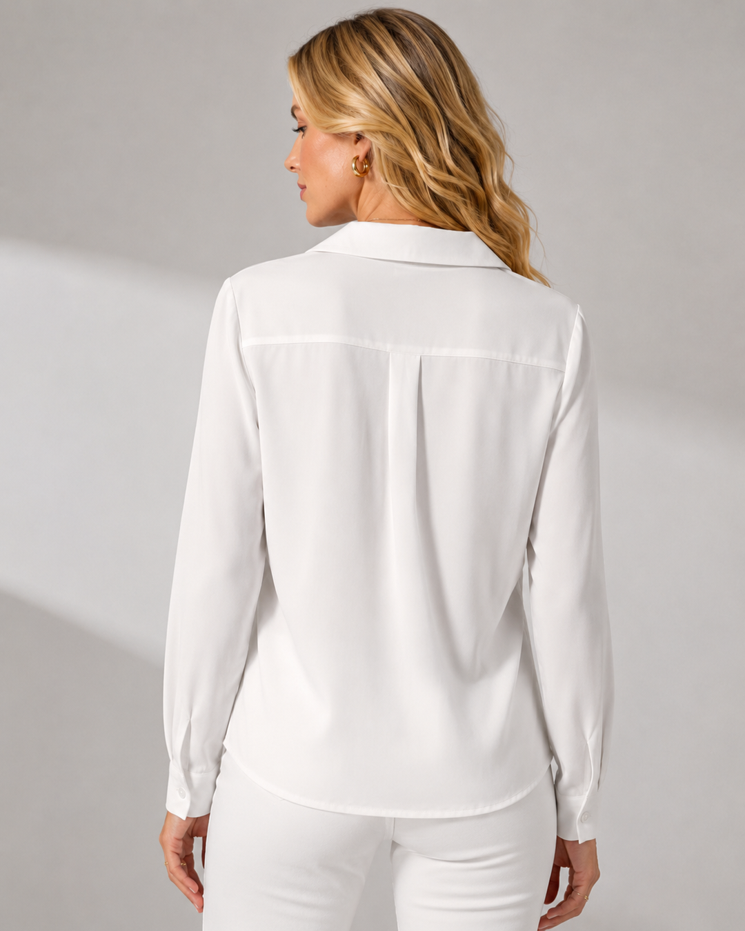 A woman shown from the back wearing a Boss lady M white long-sleeve collared blouse with a subtle pleated detail at the center back, paired with white pants, against a light gray studio background.
