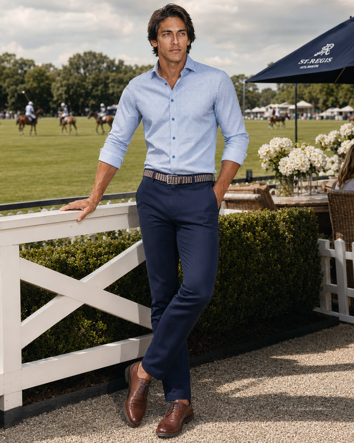 A man stands outdoors at an equestrian race course wearing a light blue long-sleeve button-down Square Up shirt, navy Venture trousers, a patterned belt, and brown leather shoes. A white fence, manicured hedges, and a blue Scalo umbrella are visible in the background.
