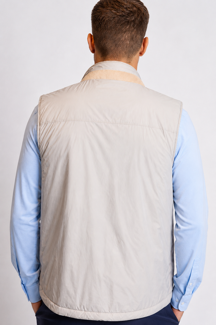 A man viewed from behind wearing a Buki light beige quilted vest over a Single Shot in Ice Flow light blue dress shirt, against a plain light gray studio background.
