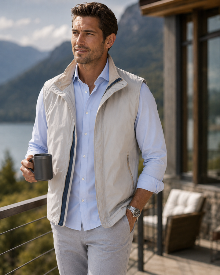 A man wearing a light beige sleeveless zip-front Buki vest over a Single Shot light blue button-up shirt and light gray pants, standing on a balcony with a mountain and lake in the background and holding a dark gray mug.