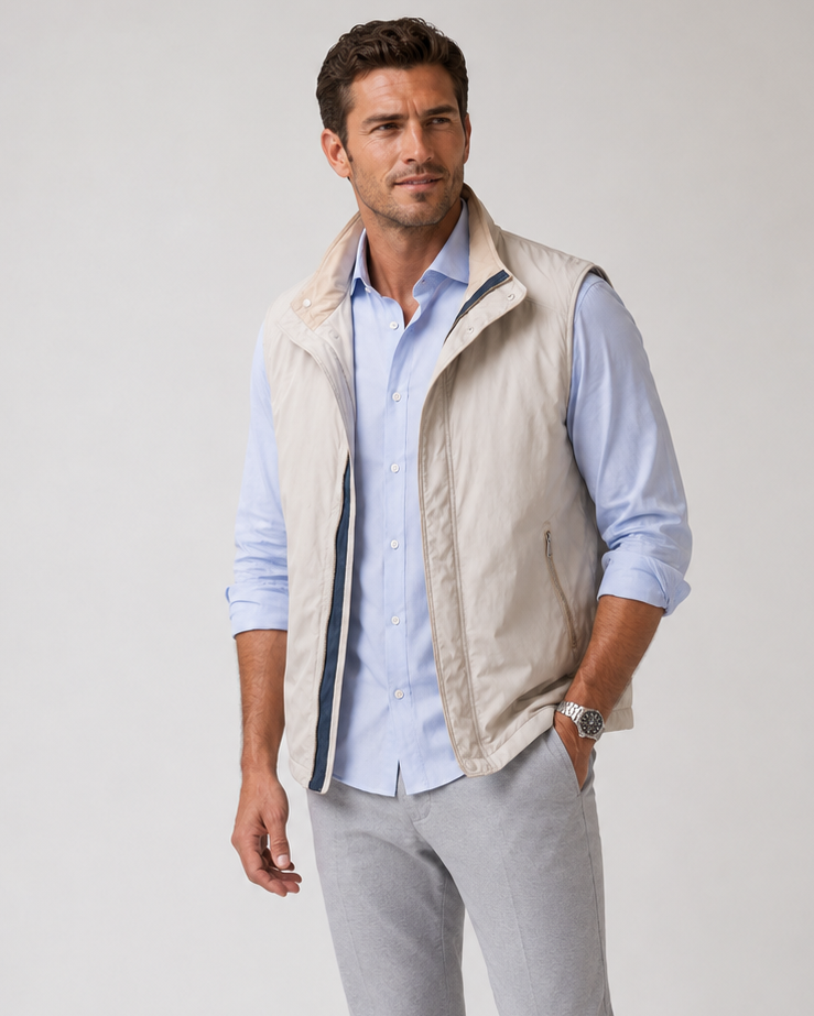 A man modeling a light beige Buki zip-up vest over a Single Shot light blue button-down shirt, paired with light gray trousers and beige loafers, against a plain light gray studio background.