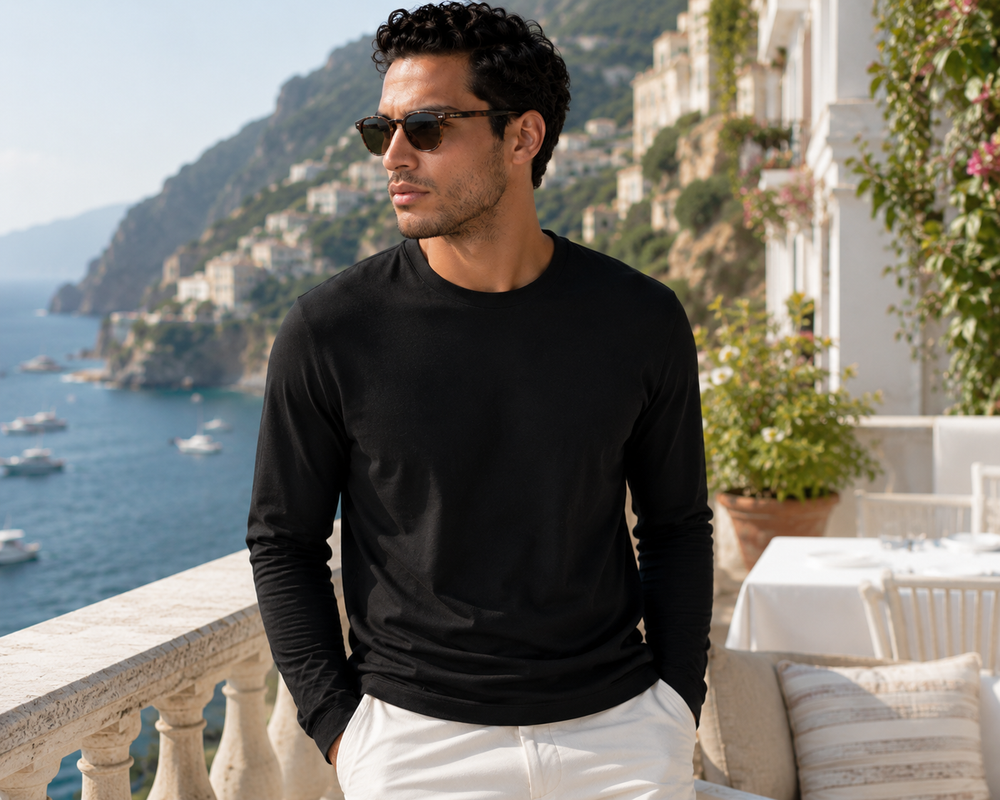 Man wearing sunglasses and a black long-sleeve Buki Cinqo shirt standing on a balcony with a scenic view of the sea and buildings in Italy.