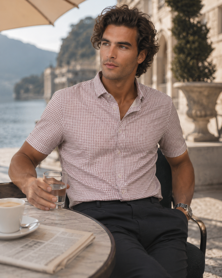 A man sitting at an outdoor patio table wearing a short-sleeve button-up Livewire shirt with a fine pink and white gingham check pattern, paired with dark trousers.