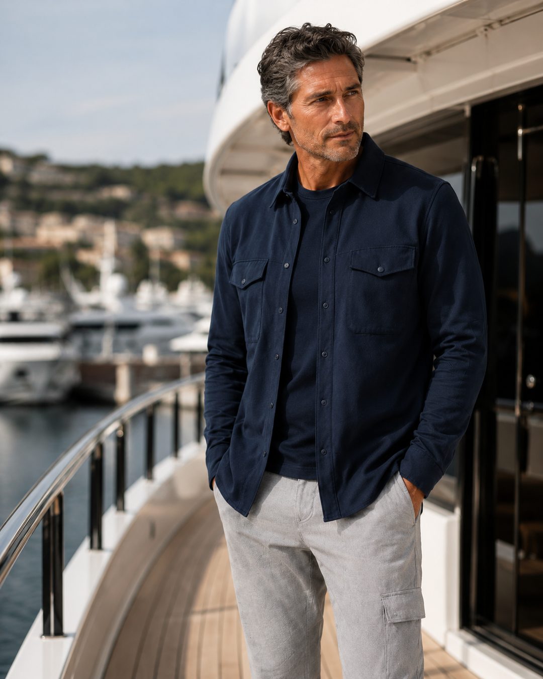 A man stands on a yacht deck wearing a Buki Atlas Overshirt in navy bBuki Navy t-shirt and light gray cargo pants, with a marina and hills in the background.