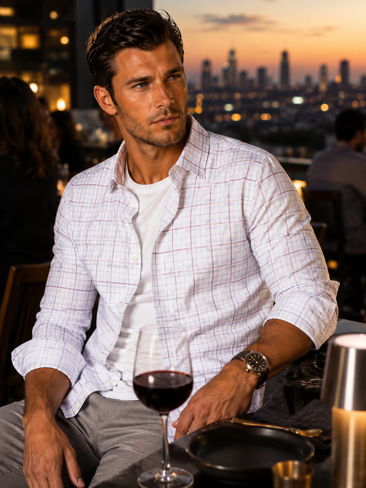 A man wearing a white long-sleeve button-down Matt Plaid shirt with a subtle pink grid pattern, layered over a white Cinqo t-shirt, sitting at a table with a glass of red wine in front of a city skyline at sunset.