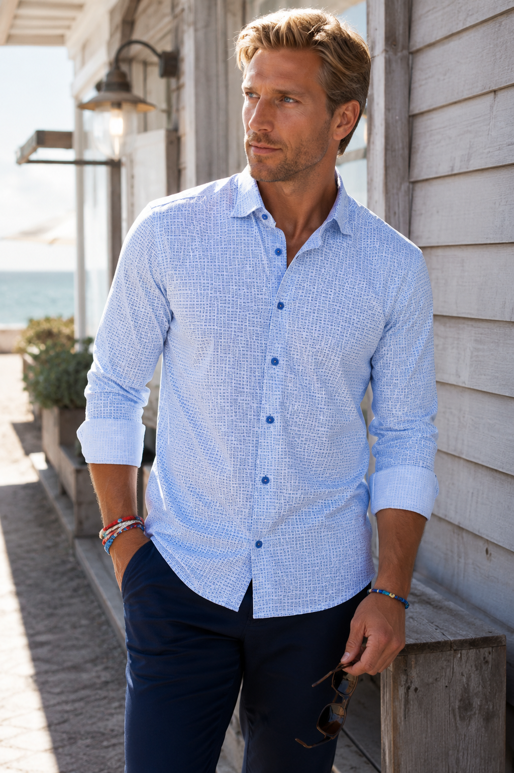 A man wearing a Ride the Wave Buki Long Sleeve Button down Tech shirt L with dark navy pants, standing outdoors against a white wooden building.