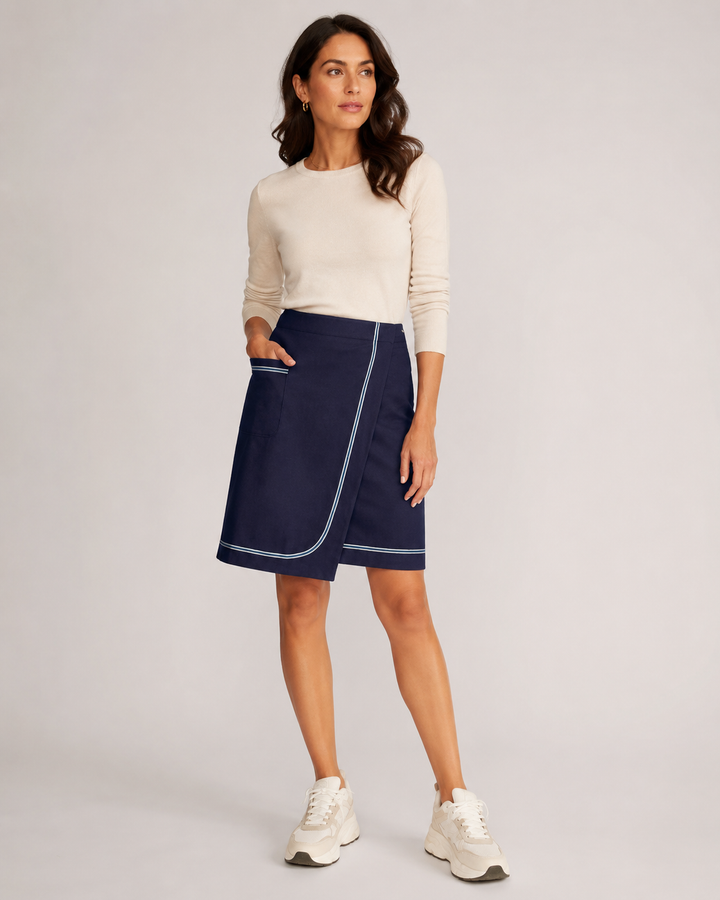 A woman modeling a Riviera skirt with light blue piping and side pockets, paired with a cream long-sleeve top and white sneakers, against a light gray studio background.