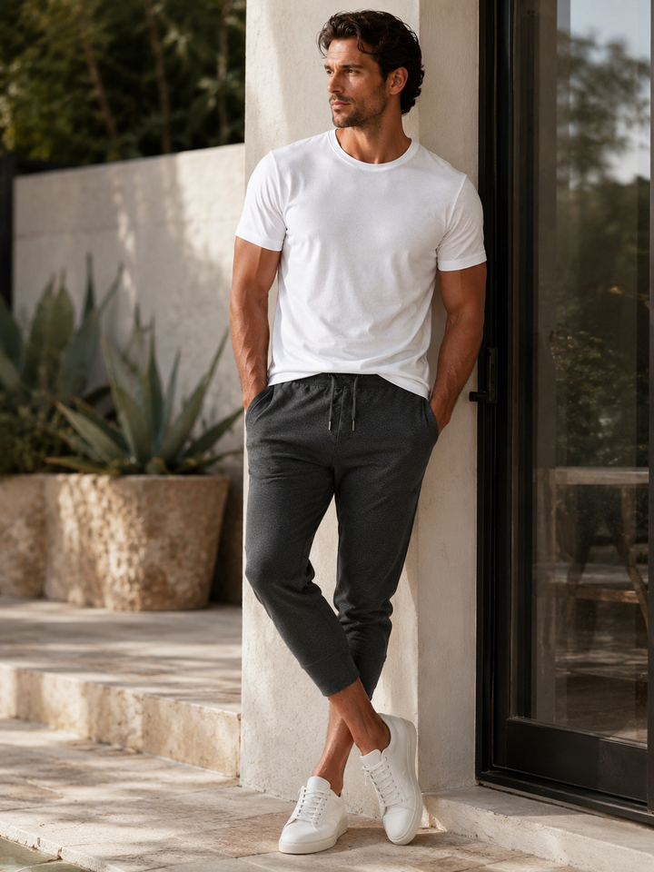 A man wearing a white Cinqo crewneck t-shirt, dark gray 
Scrimmage drawstring jogger pants L, and white sneakers, leaning against a beige stone pillar next to a glass door.