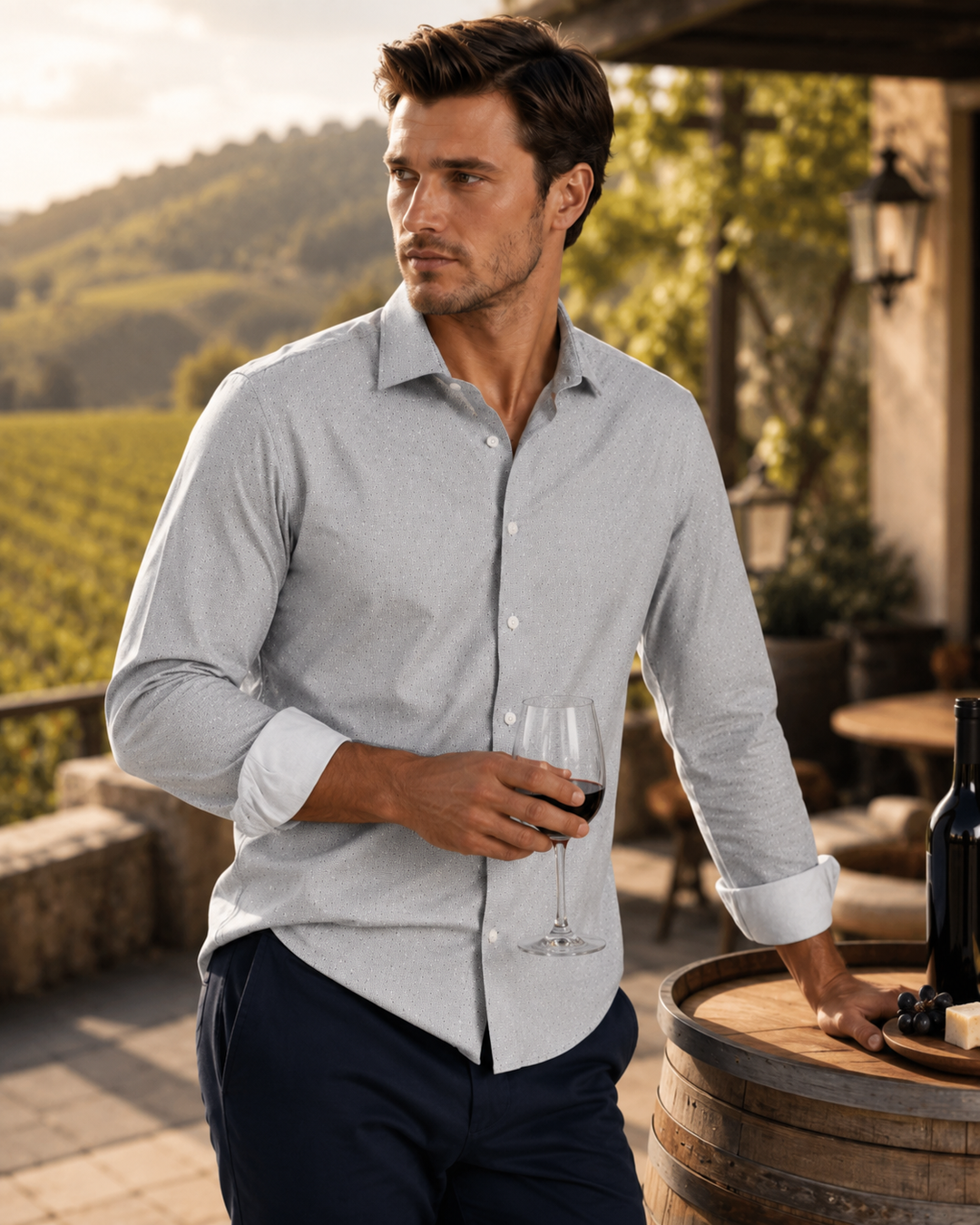 A man wearing a Studio Dot Silver long-sleeve button-down dress shirt and navy trousers stands on a vineyard terrace holding a glass of red wine, with a wooden barrel and wine bottle nearby.