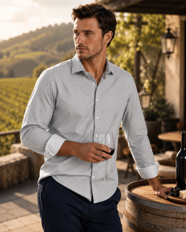 A man wearing a Studio Dot Silver long-sleeve button-down dress shirt and navy trousers stands on a vineyard terrace holding a glass of red wine, with a wooden barrel and wine bottle nearby.