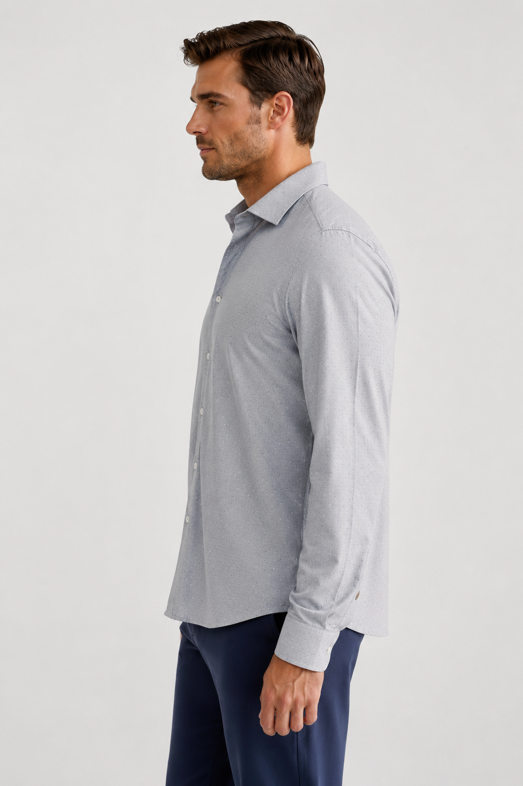 A man in side profile wearing a Studio Dot Silver long-sleeve button-down shirt and navy trousers, photographed against a plain light gray studio background.