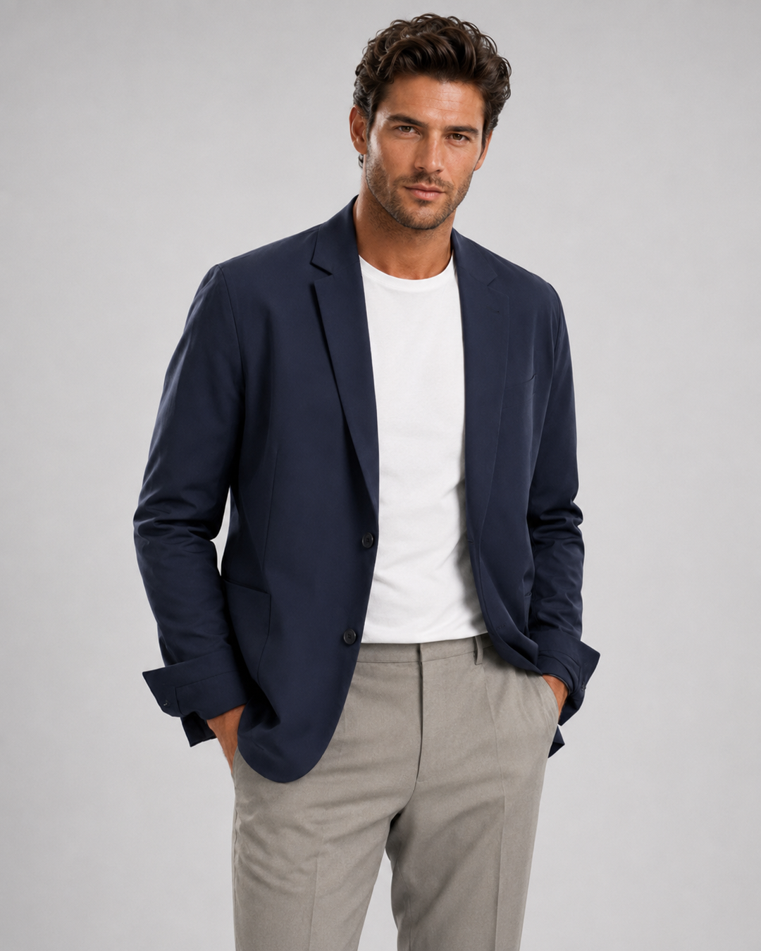 A man wearing a Buki blue travel blazer over a white t-shirt and light gray trousers, standing against a solid gray studio background.