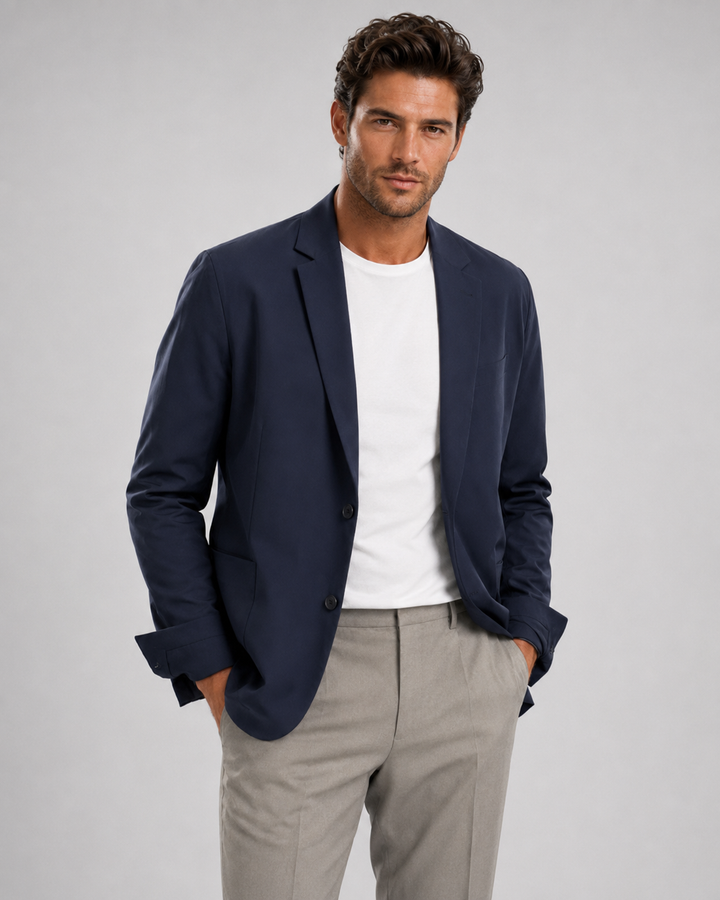 A man wearing a Buki blue travel blazer over a white t-shirt and light gray trousers, standing against a solid gray studio background.